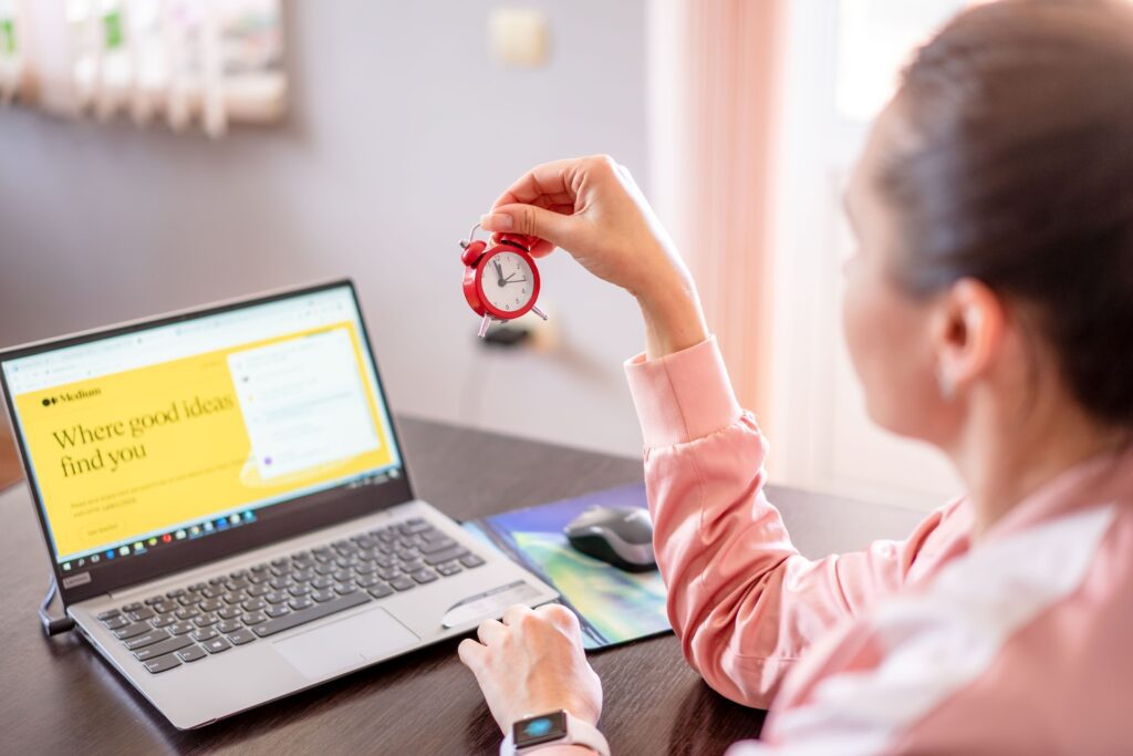 A woman working on her notebook while staring at a small red clock.